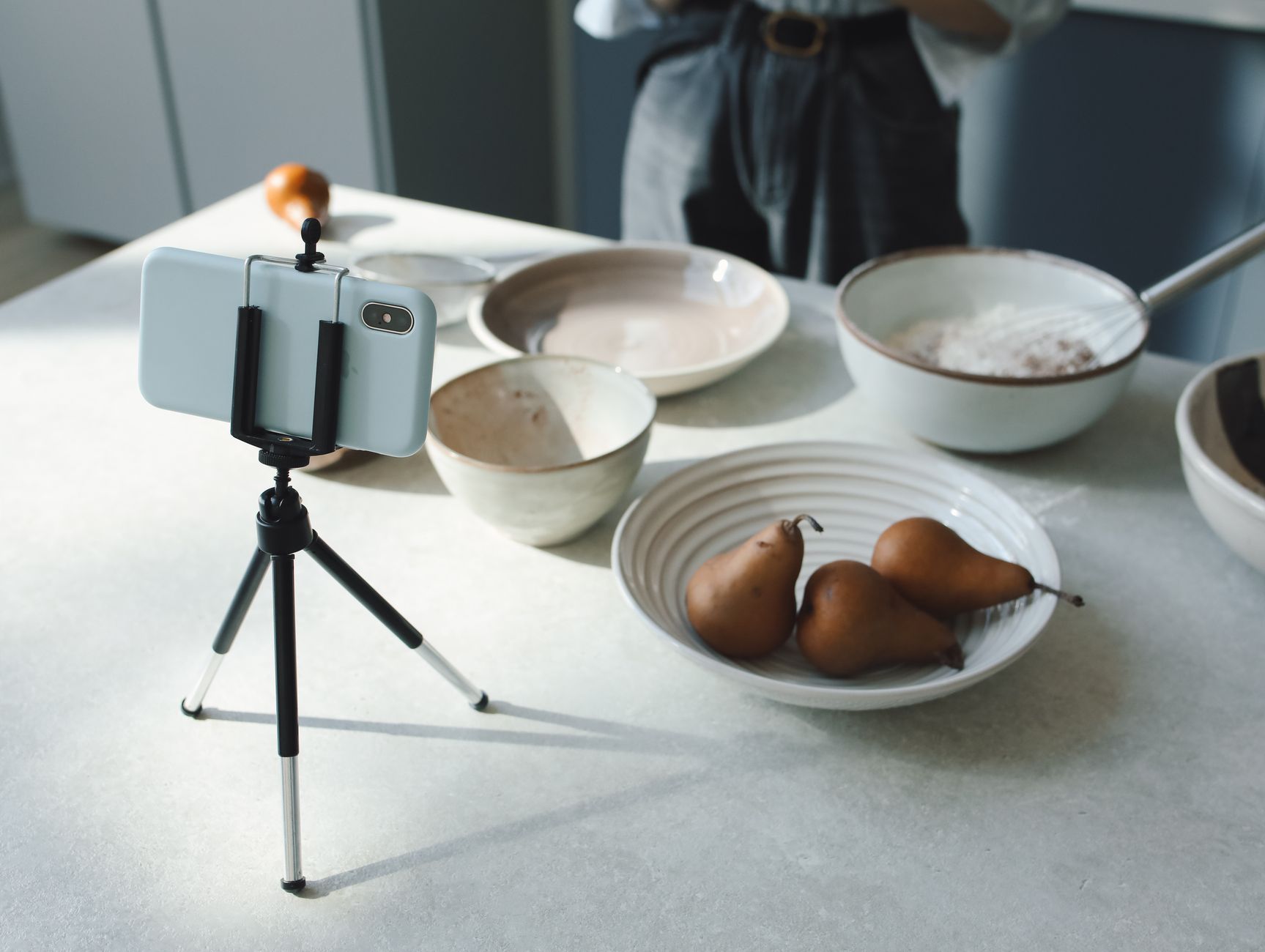 Een smartphone op een klein statief op een keukentafel met keramische kommen en peren